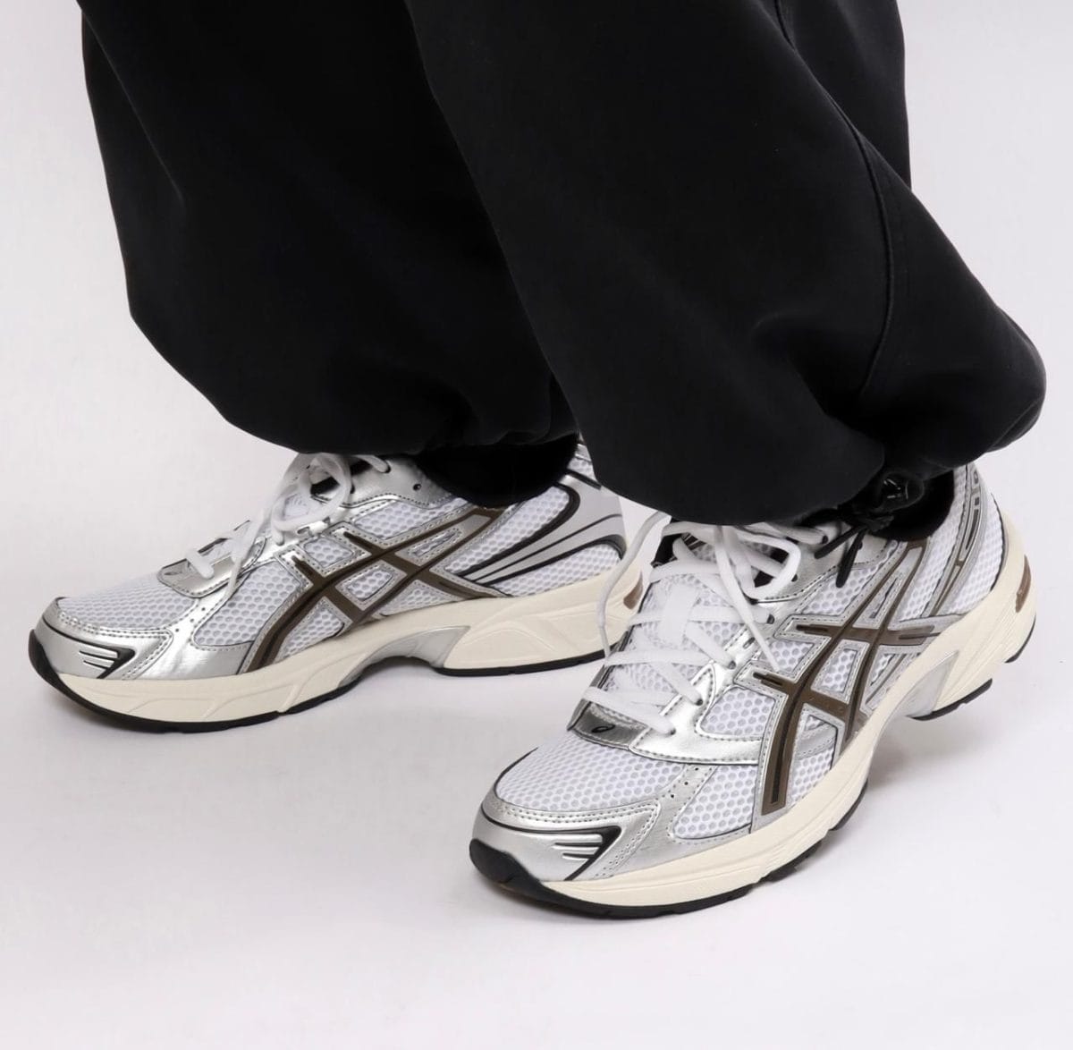 Close-up of feet in white and silver running shoes with mesh uppers, standing on a white surface while wearing black pants.