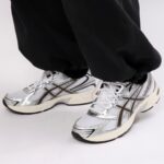 Close-up of feet in white and silver running shoes with mesh uppers, standing on a white surface while wearing black pants.