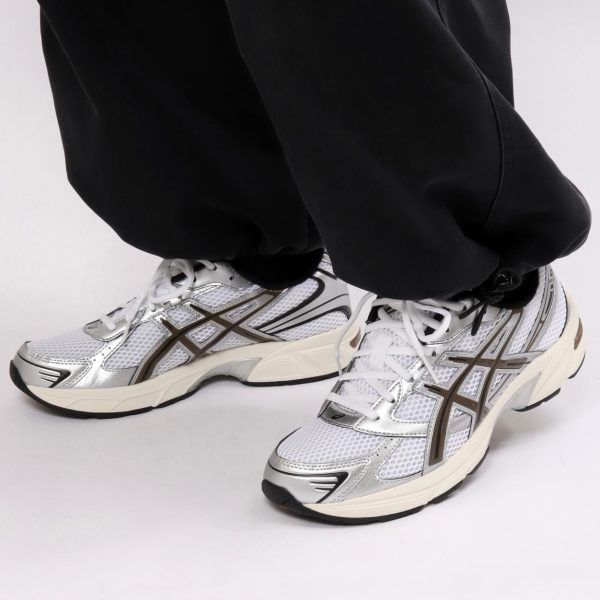 Close-up of feet in white and silver running shoes with mesh uppers, standing on a white surface while wearing black pants.