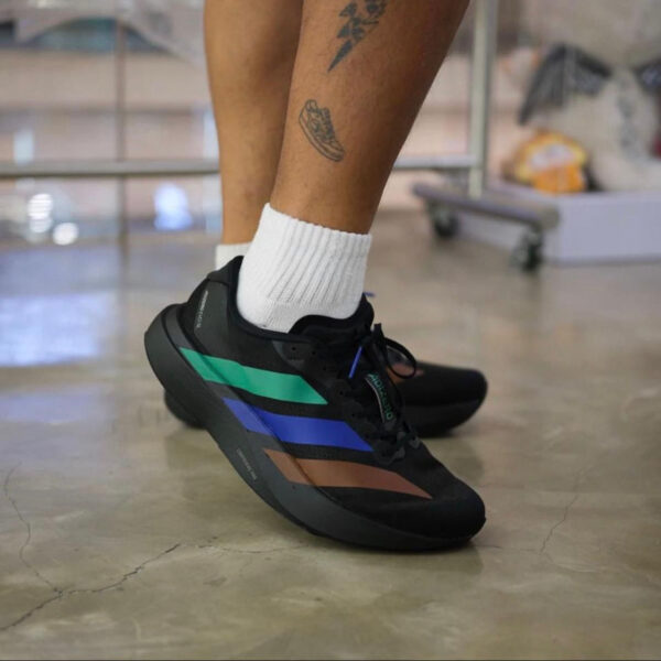 Close-up of a person wearing black sneakers with green, teal, blue, and brown stripes and white socks on a polished floor.