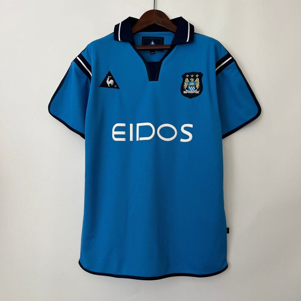 Blue soccer jersey on a wooden hanger, featuring the Le Coq Sportif logo on the left chest and the club crest on the right, with large white lettering across the front.