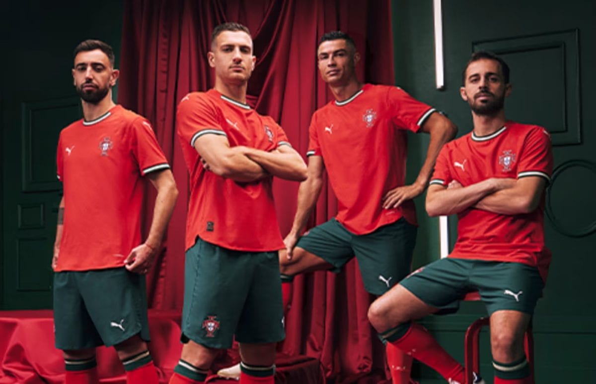 Four male soccer players in red jerseys and green shorts pose together indoors.