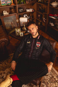 Man lounging in a leather chair in a cozy room, wearing a black pinstripe sports outfit with a crest, looking at the camera.