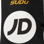 Close-up of black patterned fabric with an orange 'SDU' logo above a large white circle containing a black letter 'D'. The image shows branding detail on a textile item.