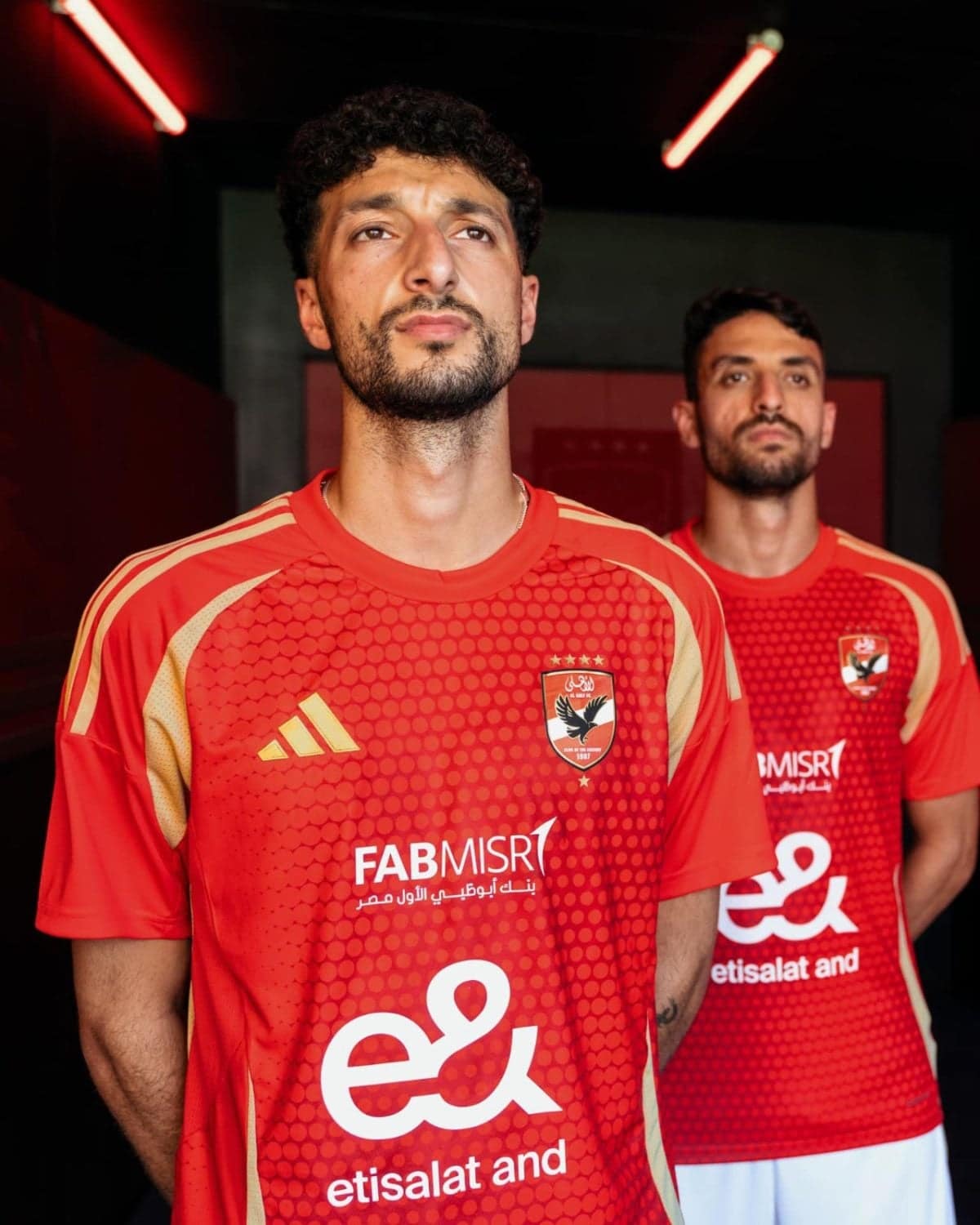 Two male soccer players in red jerseys with team crest, standing in a dim hallway with red lighting; sponsor logos visible on front.
