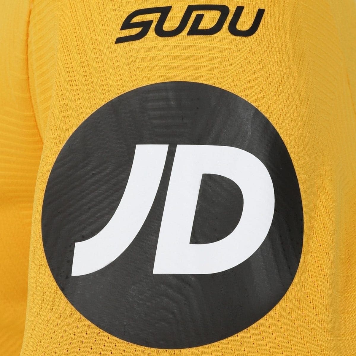 Close-up of bright yellow knit fabric with a large black circular patch bearing a white stylized 'JD' logo and partial 'SUDU' text above.