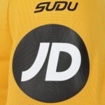 Close-up of bright yellow knit fabric with a large black circular patch bearing a white stylized 'JD' logo and partial 'SUDU' text above.