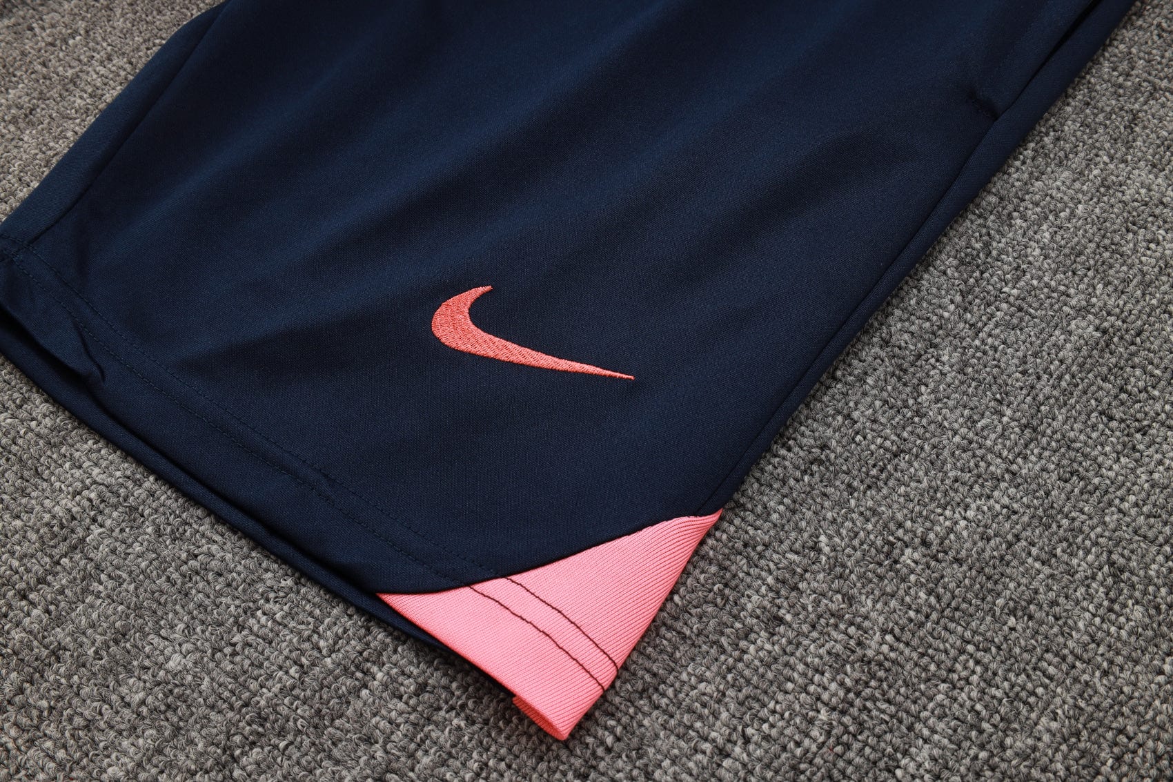 ca5c12a7 | UAE SPORT STORE Close-up of navy athletic pants with a pink embroidered Nike swoosh and pink hem resting on gray carpet.