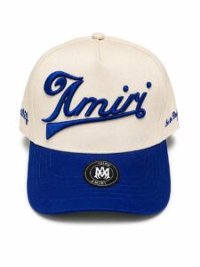 Beige hat with blue embroidered 'Amiri' logo across the front and a blue curved brim with a circular logo sticker on the bill.