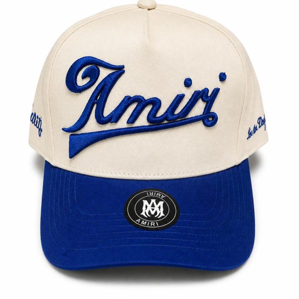 Beige hat with blue embroidered 'Amiri' logo across the front and a blue curved brim with a circular logo sticker on the bill.