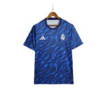 Blue Real Madrid football jersey with a wavy pattern, crest on the left chest and Adidas logo on the right, hanging on a wooden hanger.