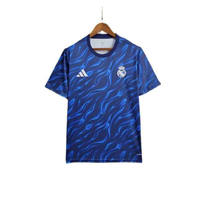 camisa-treino-do-real-madrid-adidas-2025-2026-treinamento-academia-rodrygo-azul-vini-jr-torcedor-masculina-homem-torcedor-original-oficial-comprar-online-frete-grati-4f14a61875d4c13b7 Blue Real Madrid football jersey with a wavy pattern, crest on the left chest and Adidas logo on the right, hanging on a wooden hanger.