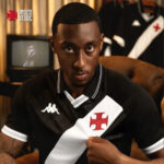 Portrait of a man in a black-and-white diagonal-split soccer jersey with a red cross crest, seated and looking intently at the camera in a cozy room.
