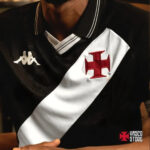 Close-up of a person wearing a black Vasco da Gama soccer jersey with a white diagonal sash and a red cross emblem, and a small Kappa logo on the left.