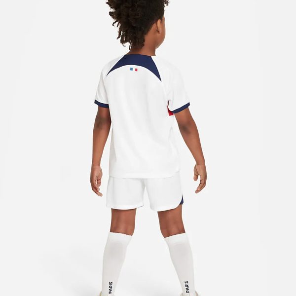 Child in white soccer kit seen from the back with navy accents, white shorts, knee-high socks that read PARIS, and colorful soccer cleats.