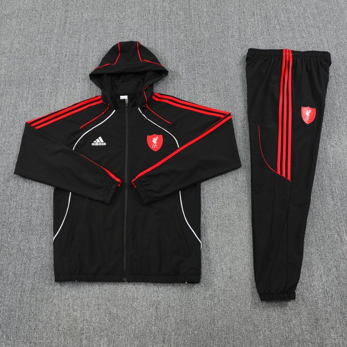 Liverpool FC black training tracksuit laid flat on a gray carpet, jacket with red piping and crest, matching pants with red stripes.