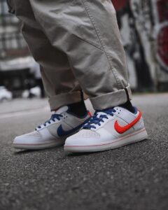 Close-up of white Nike sneakers with blue laces and red swoosh, worn with rolled-up gray pants, standing on pavement.