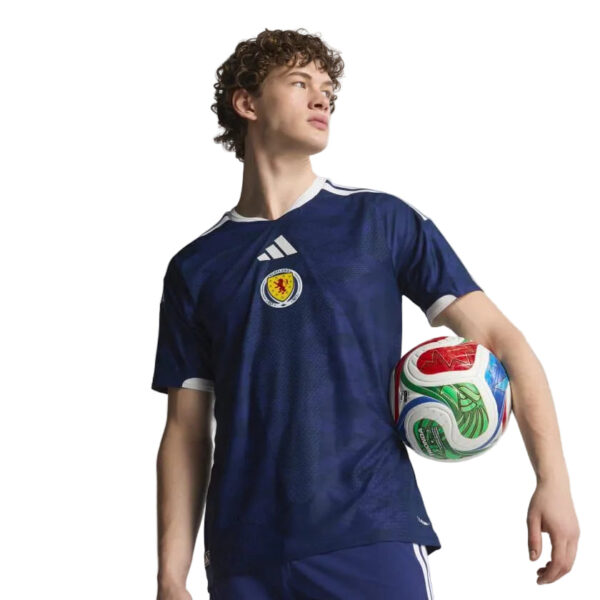 Young man in a navy soccer jersey holds a colorful soccer ball under one arm, looking off to the side against a white background.