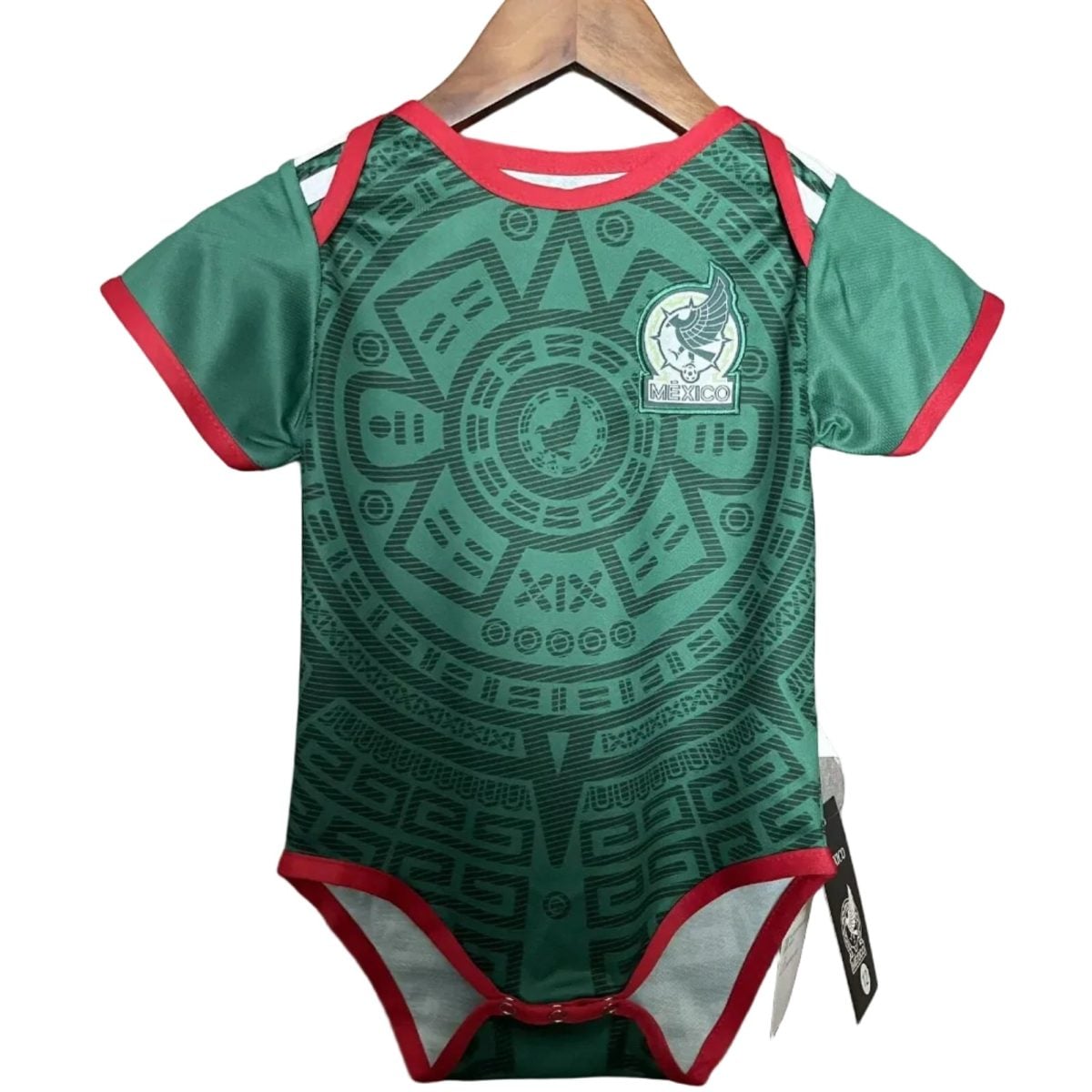 Green baby onesie with red trim and an intricate circular pattern, featuring a Mexico crest with an eagle on the chest, hanging on a wooden hanger.