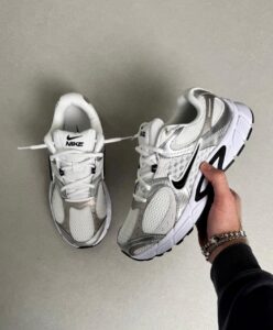 Two white Nike sneakers on a light gray floor; one is held by a hand with bracelets, the other sits nearby.