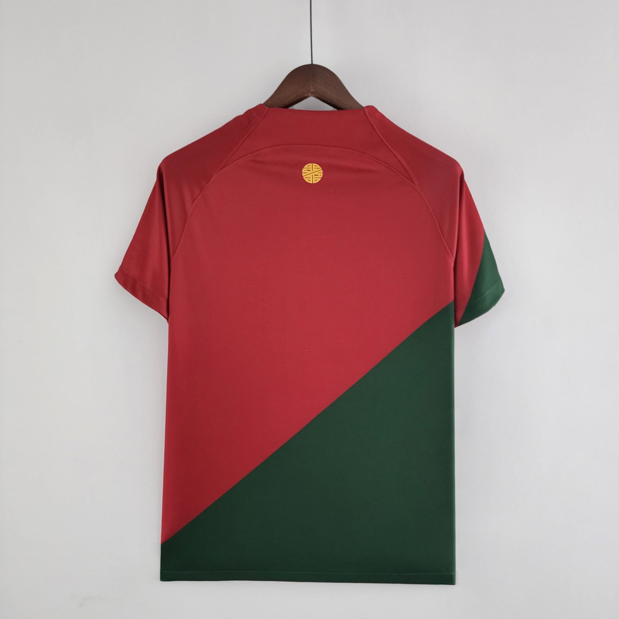 cdc063d5_9b5d5c2c-01ac-4f01-90b8-613ce590c041 | UAE SPORT STORE Back view of a red shirt on a wooden hanger, featuring a small yellow circular logo near the neckline.