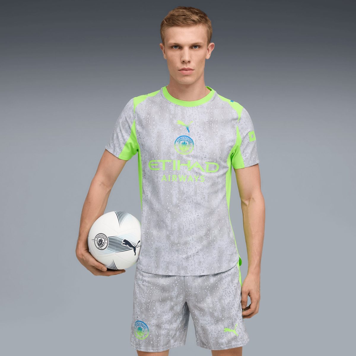 Young male soccer player in a gray Manchester City jersey with neon green accents, holding a white soccer ball against a gray backdrop