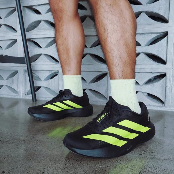 Person wearing black running shoes with bright neon-yellow stripes and white socks, standing on a concrete floor beside a chair.