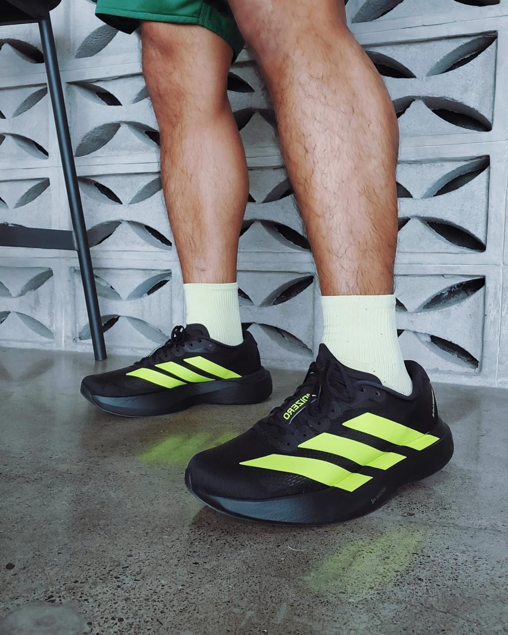 ce31a2dd-fff2-426a-8077-4ba28e72d12f | UAE SPORT STORE Person wearing black running shoes with bright neon-yellow stripes and white socks, standing on a concrete floor beside a chair.