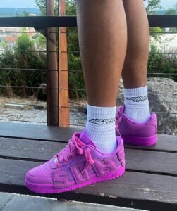 Person standing on a wooden deck wearing white socks and bright pink sneakers, outdoors with greenery in the background.