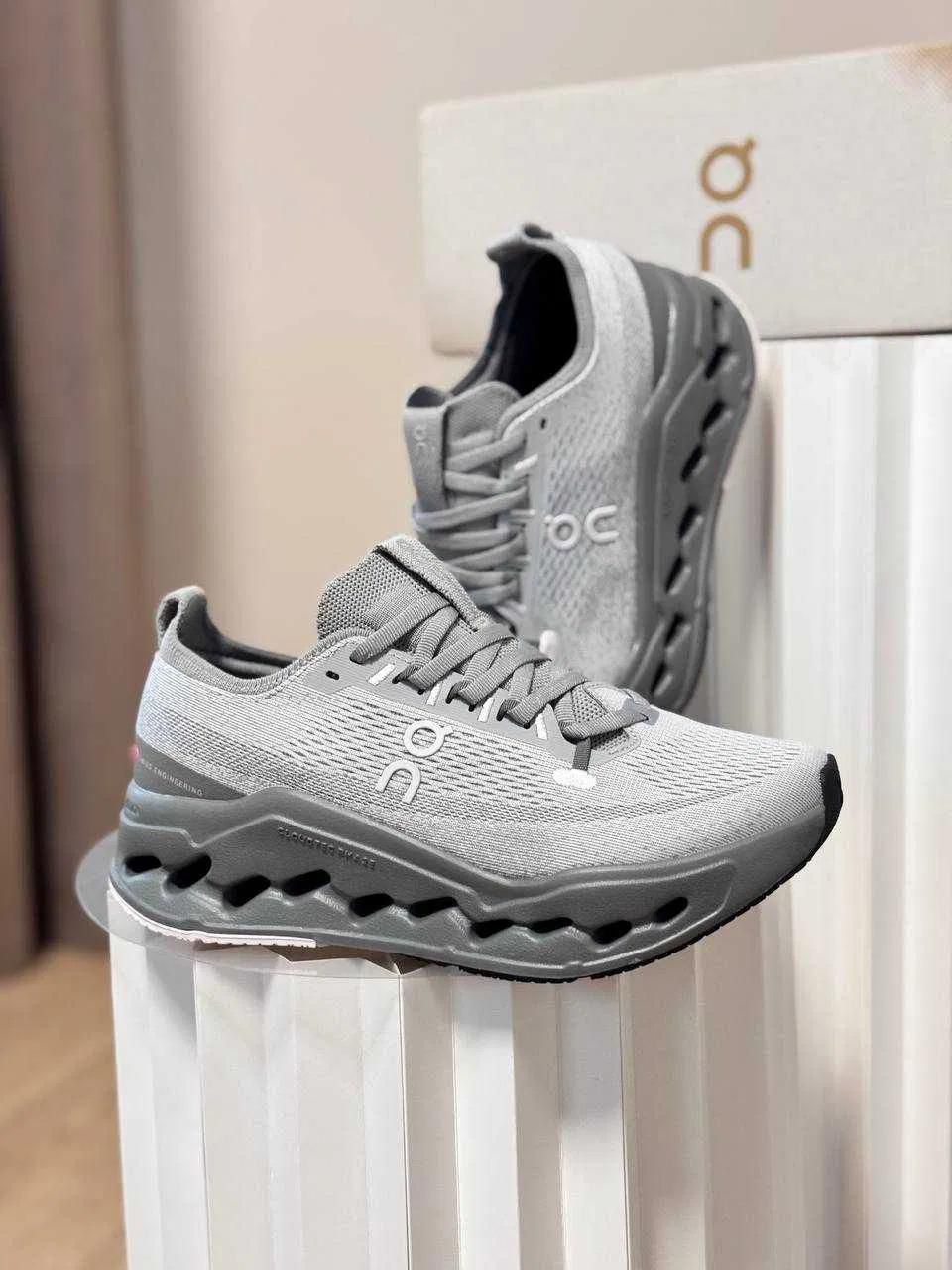 cef2c425-6f2e-4e74-80a0-ddcd43598fbb | UAE SPORT STORE Pair of gray running shoes with knit upper and sculpted midsole on a white display pedestal in a store, showing the ON logo on the side.