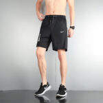 Male model wearing black Nike gym shorts and black sneakers, standing with one hand on hip.