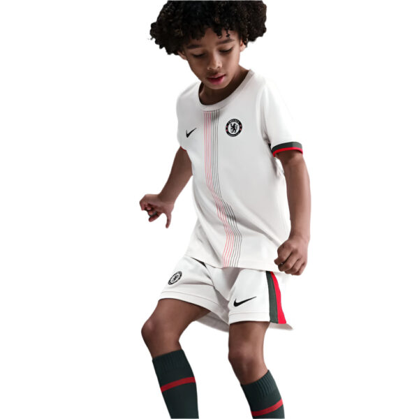 Young boy in a white Chelsea FC youth soccer kit (jersey and shorts) with Nike logos and red/green trim, mid-action pose with dark socks.