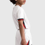 Side view of a person wearing a white sports jersey with black and red trim on the sleeve and side paneling, elastic waistband shorts, neutral gray background.