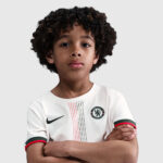Young boy with curly hair stands with arms crossed, wearing a white Chelsea FC soccer jersey with Nike swoosh.