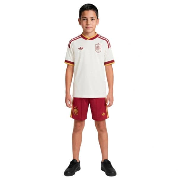 Boy wearing a Spain national team soccer jersey (white with maroon trim) and red shorts, standing in black sneakers against a white background.