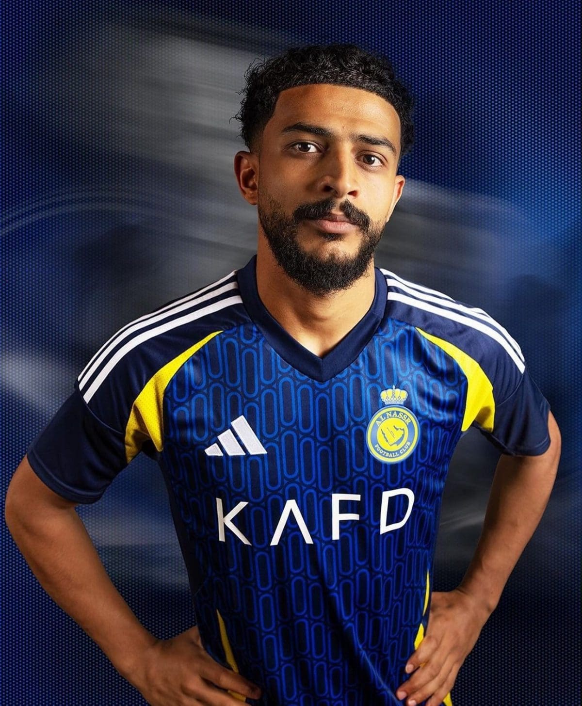 Portrait of a man in a blue football jersey with yellow accents, hands on hips, posing against a dark blue backdrop.