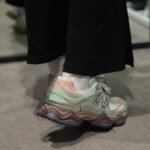 Close-up of pastel multicolor sneaker with mint green, peach, and beige tones on a gray floor, worn with white socks and black pants.