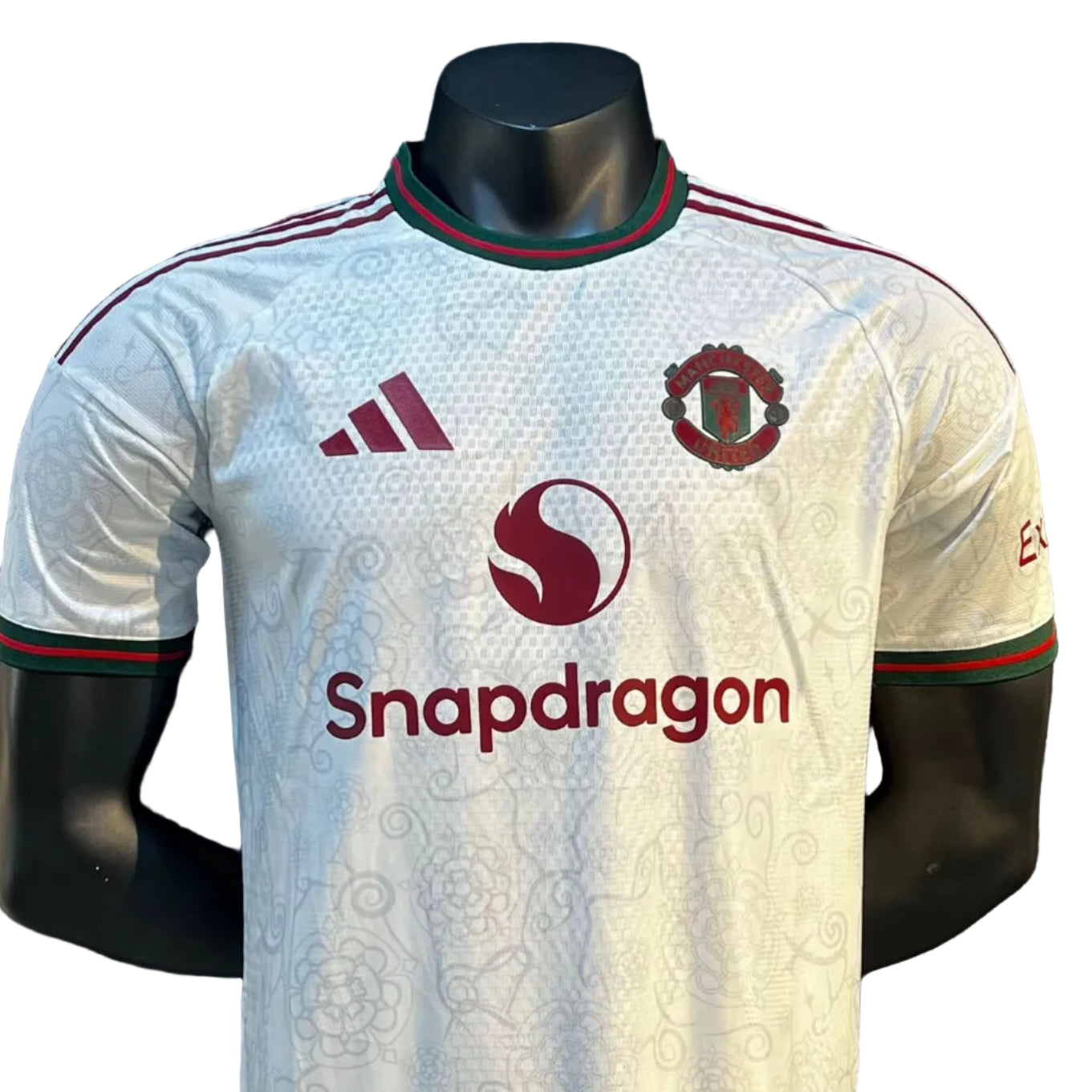 d0c1dac0-7849-44e0-9017-bf6b2b00b15e | UAE SPORT STORE Front view of a white Manchester United jersey on a mannequin, featuring the club crest, Snapdragon sponsor, and red-green trim.