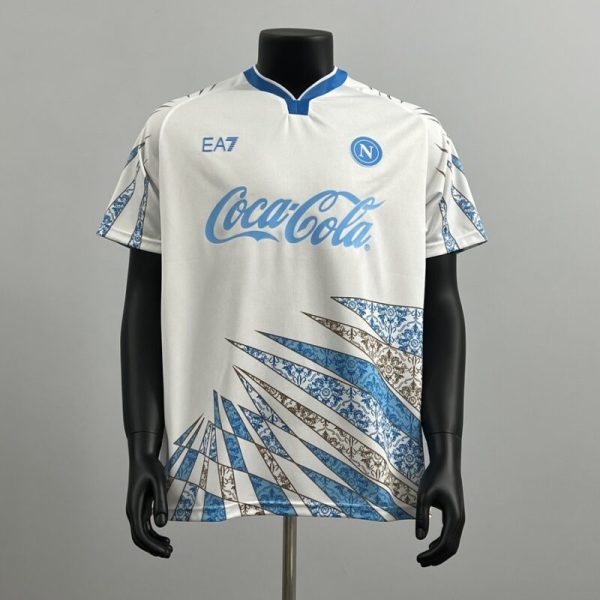White Napoli football jersey with blue Coca-Cola sponsor, Napoli crest, and blue-brown angular pattern at the lower front.