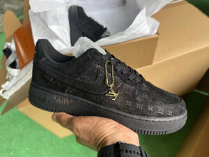 Black Louis Vuitton monogram sneaker with a gold LV charm, held in hand over a shoebox.