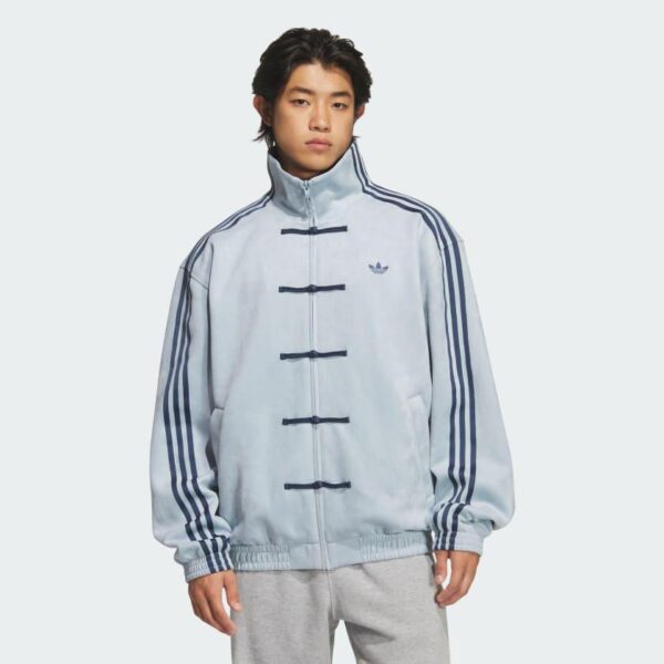 Young man wearing a light blue Adidas jacket with dark blue stripes and frog-button closures, standing against a pale background.