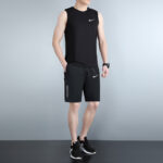 Man wearing a black Nike sleeveless top and black Nike shorts, standing with hands in pockets against a gray backdrop.