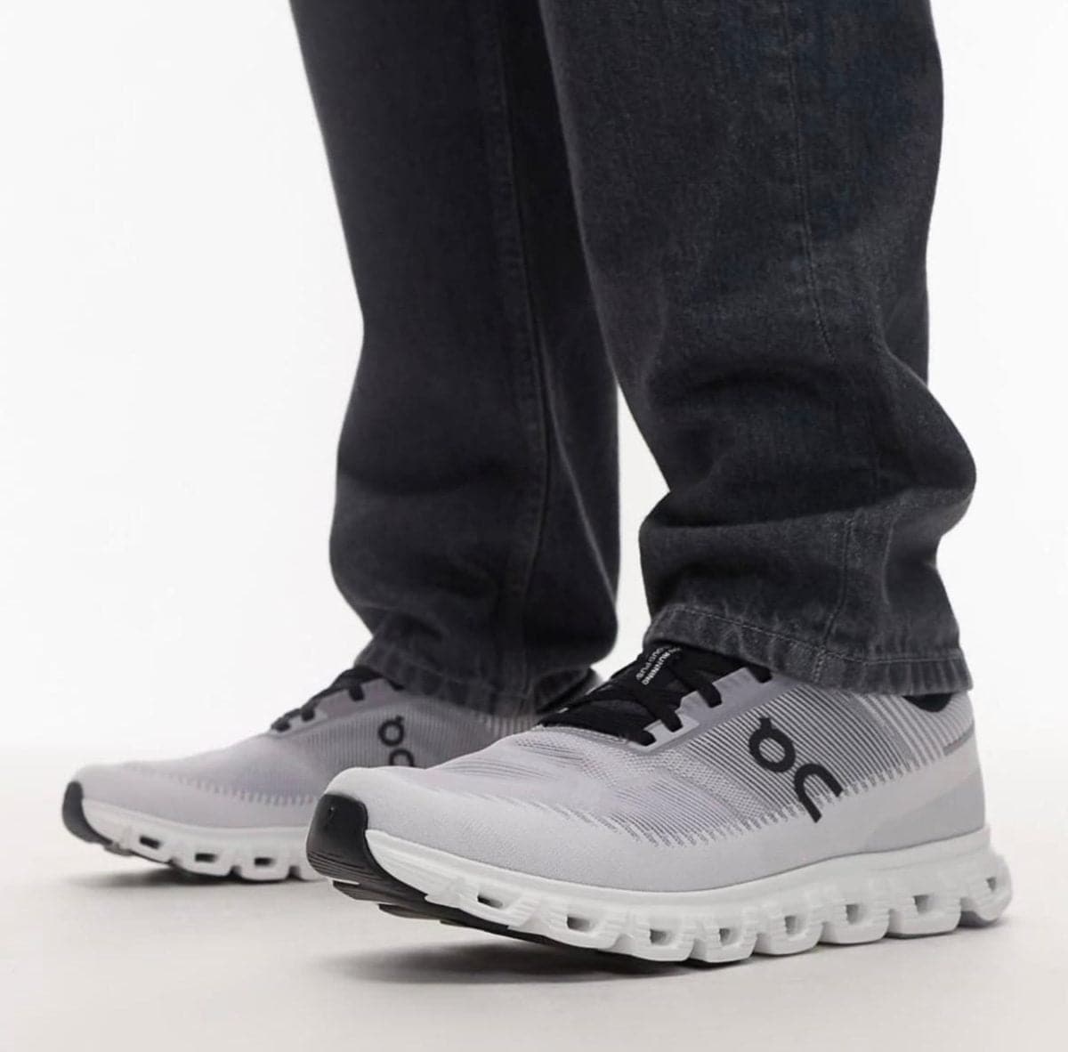 Light-gray and white running shoes with black laces worn with dark jeans, standing on a light surface.