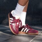 Maroon Adidas Samba sneakers with white stripes and laces, worn with white socks, standing on pavement.