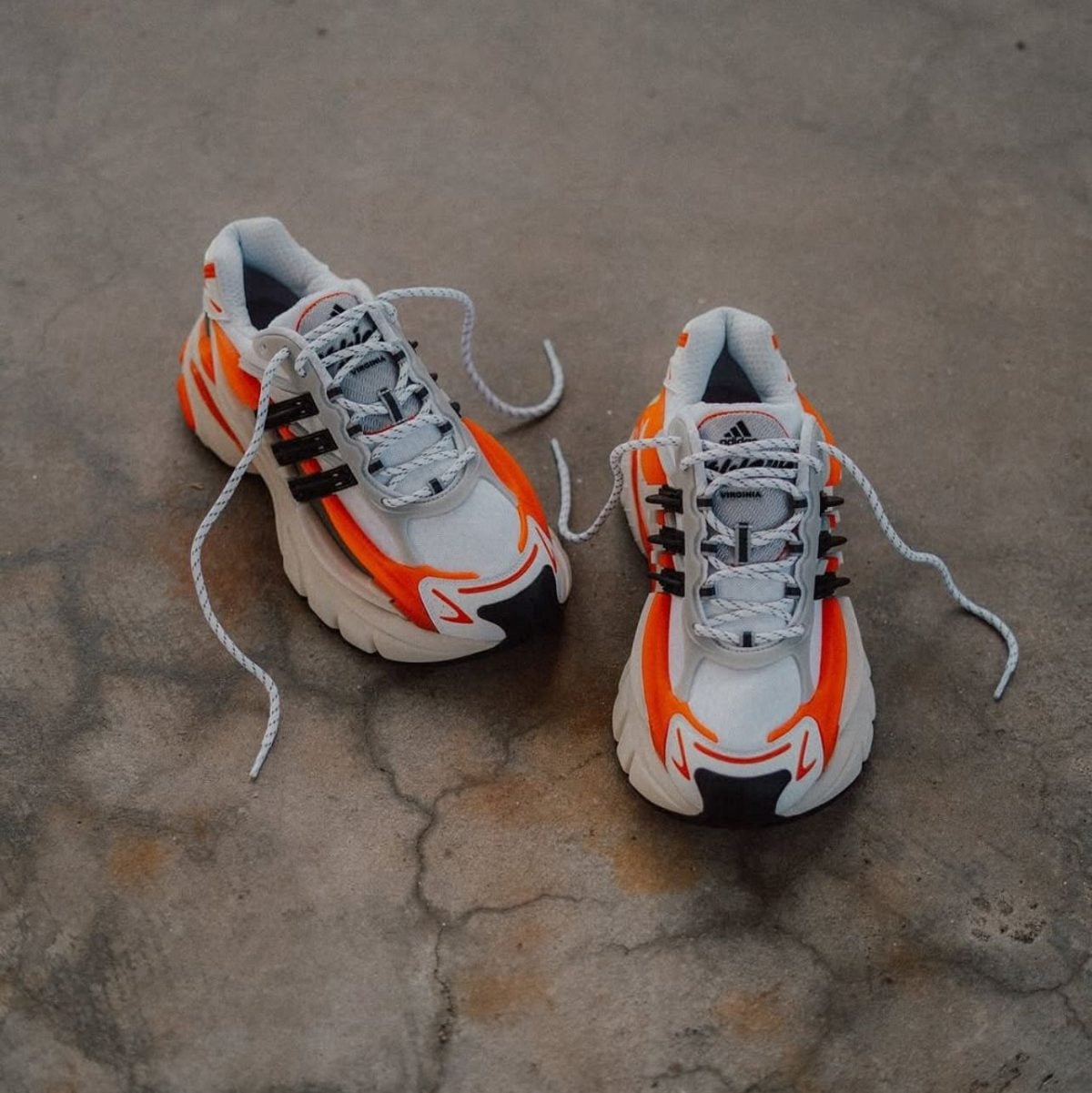 Pair of white and orange chunky sneakers on a cracked concrete floor, laces loose and hanging.