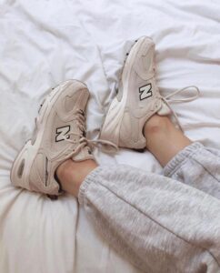 Beige New Balance sneakers with large N logo on a white bed, with legs in gray sweatpants.
