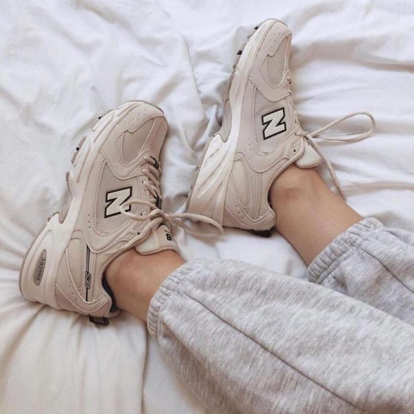 Beige New Balance sneakers with large N logo on a white bed, with legs in gray sweatpants.