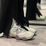 Close-up of pastel mint-green New Balance sneakers peeking from under black pants on a gray floor.