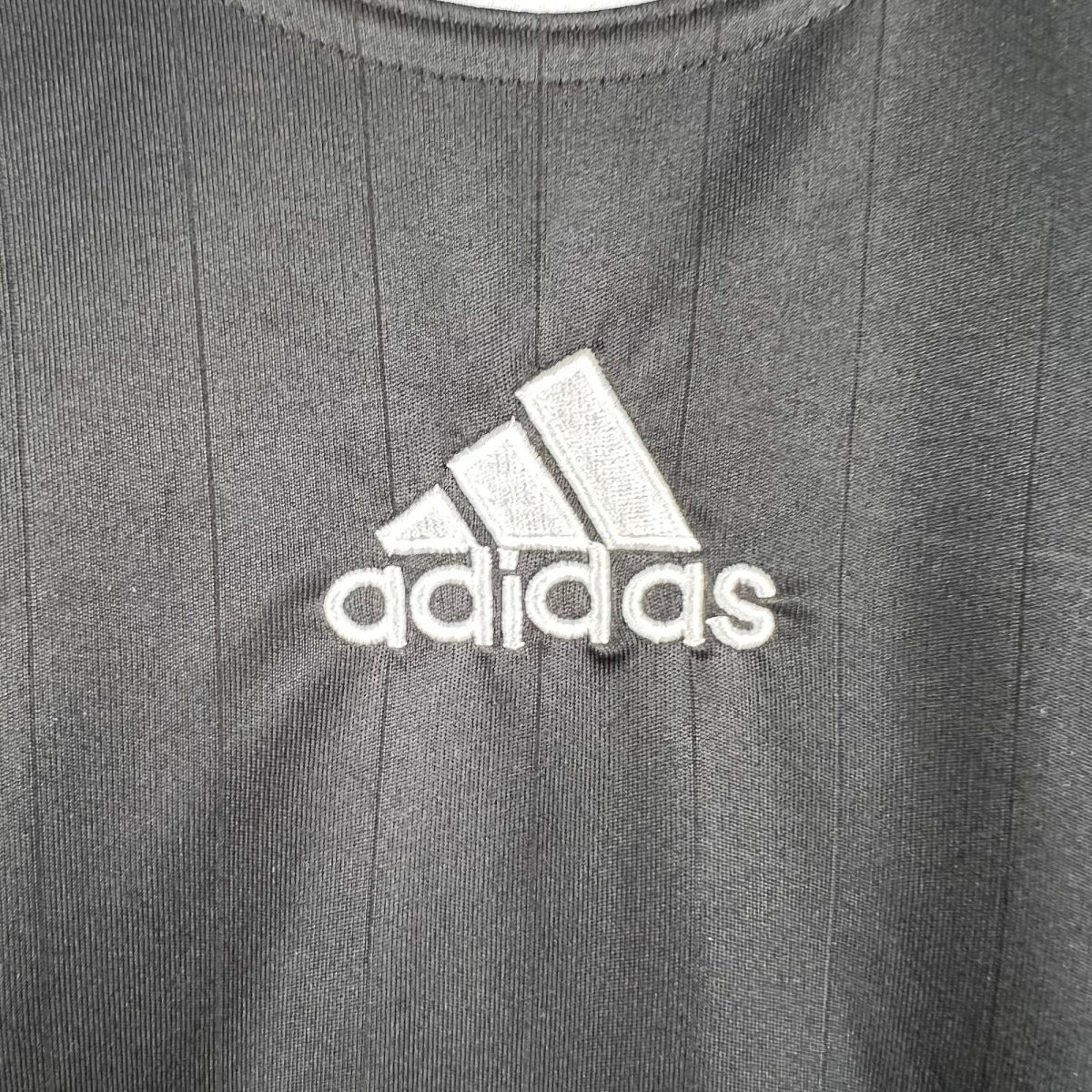 Partial view of a white Adidas logo on gray textured fabric background, with visible stitching lines around.