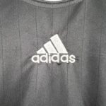 d426c6ae_af92ba70-2a8f-4965-8966-ea056bf7fbf3 | UAE SPORT STORE Partial view of a white Adidas logo on gray textured fabric background, with visible stitching lines around.